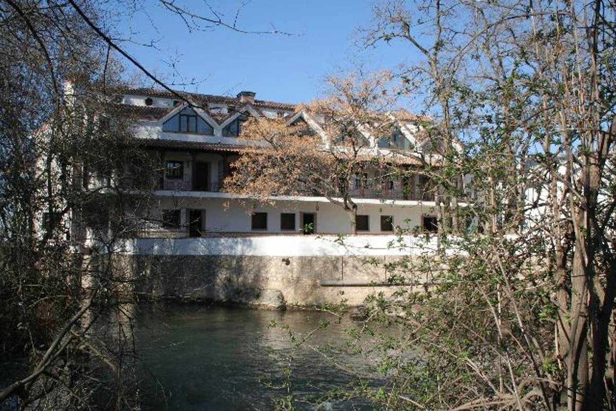 The exterior of the recently rehabilitated rural residence is presented, featuring a multi-family structure that overlooks a serene river. Trees and foliage frame the building, with large windows offering views of the surrounding nature. A sense of tranquility is conveyed by the peaceful water below.