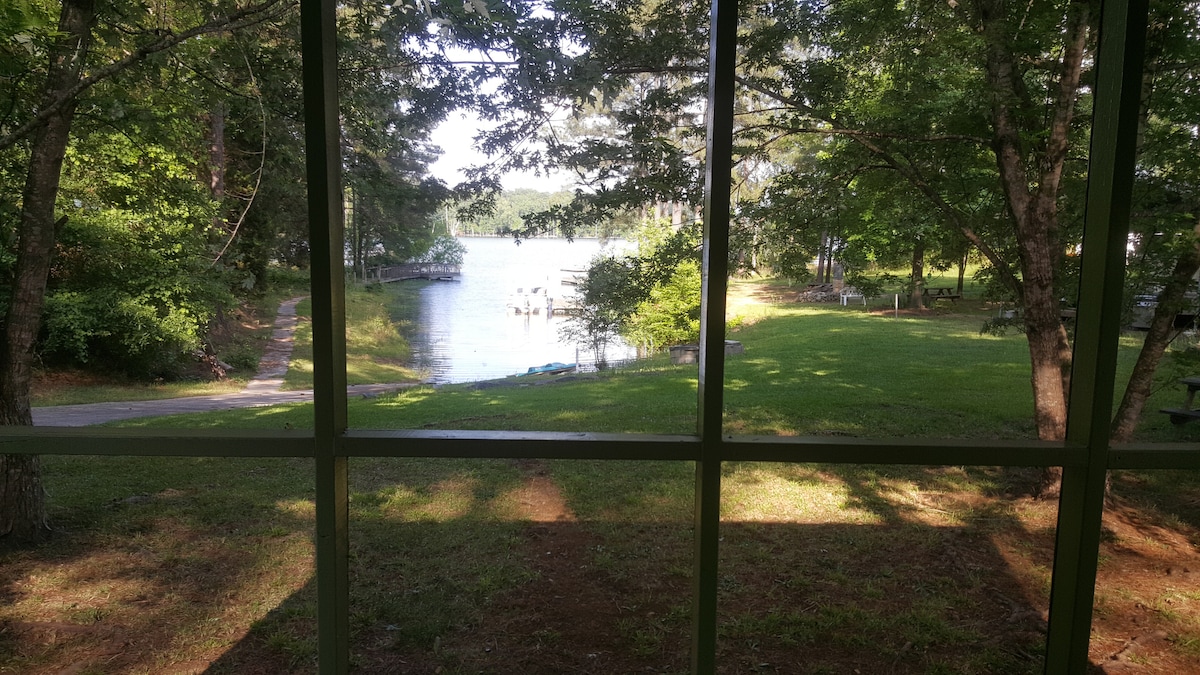 A serene view of a lake is framed by the window, showcasing the calm water and surrounding greenery. The scene includes a grassy area leading to the water, with trees providing natural shade and tranquility.