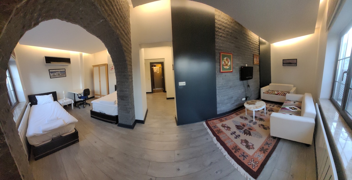 The spacious living area features a light-toned floor and stone accent walls, creating a modern setting. Two beds are visible, one with unmade sheets, and a seating area includes a small table. Large windows provide natural light, enhancing the overall openness.