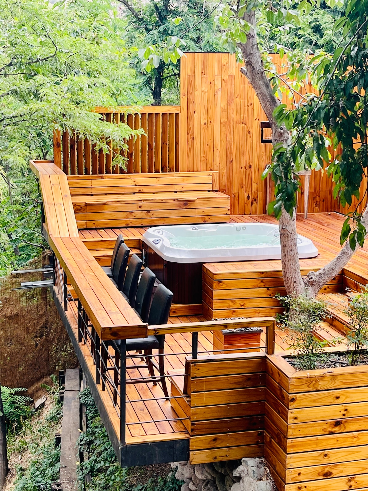 An outdoor deck features wooden paneling, with a hot tub nestled among trees. Four black chairs are positioned around a wooden table, providing a comfortable space for relaxation. The natural surroundings are visible, enhancing the feeling of seclusion.