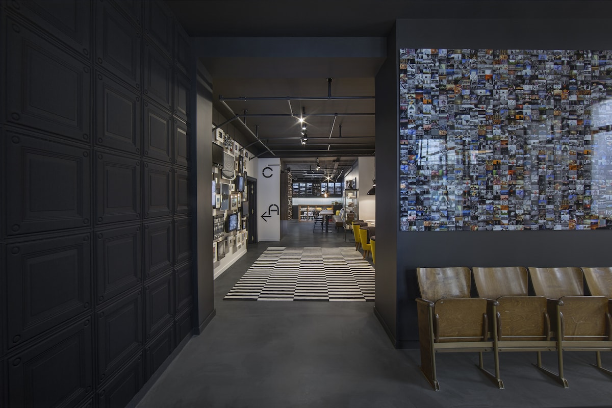 An industrial-style lobby is shown, featuring dark walls with textured paneling. A colorful interactive photo wall is present on the right, while a spacious seating area with wooden chairs and a striped rug invites relaxation. Modern lighting illuminates the entire space.
