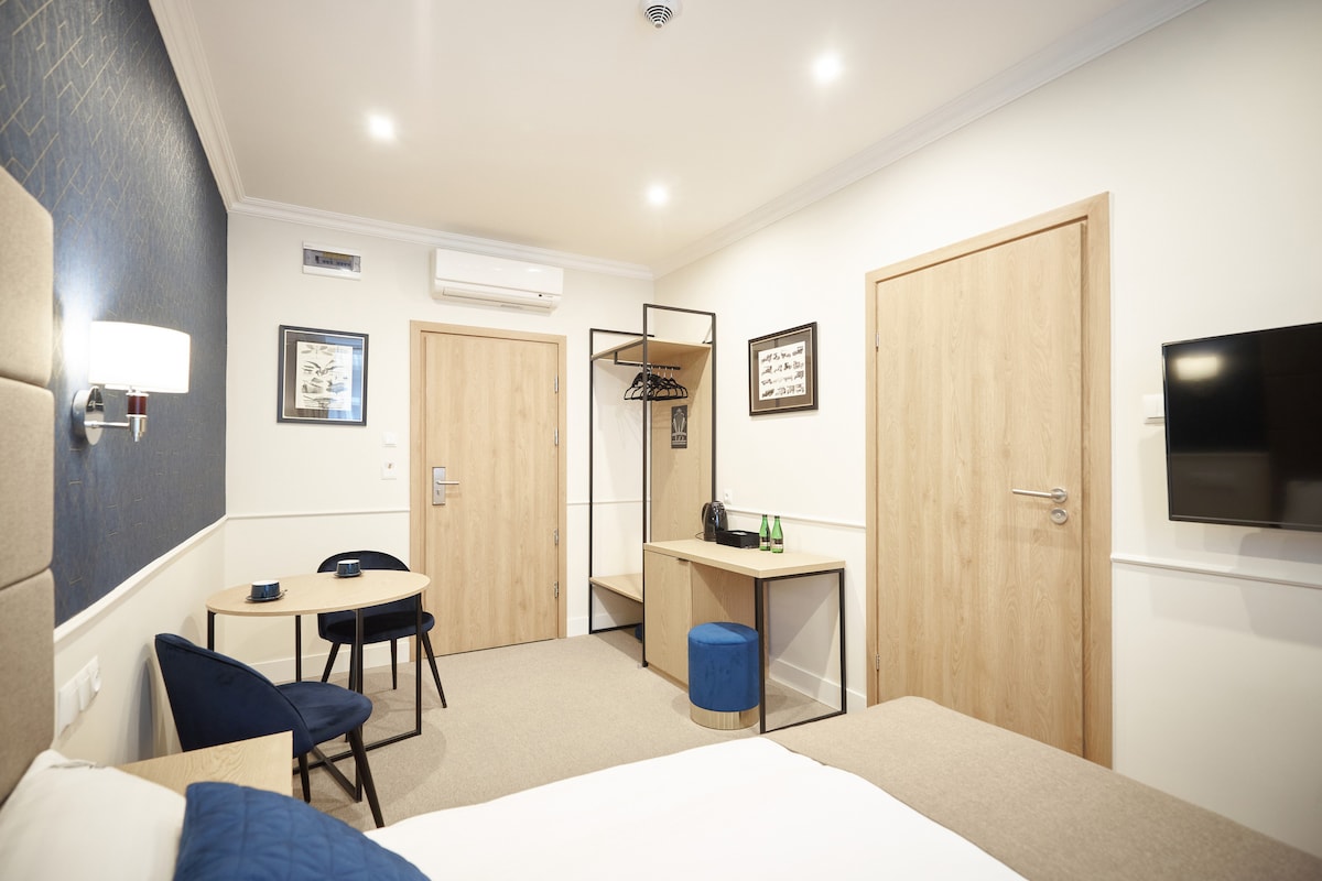 A modern guest room is displayed, featuring a double bed with a neutral color scheme. A small wooden table with two chairs and a TV are positioned against the wall. A wardrobe and a small seating area are available, complemented by a window that allows natural light.