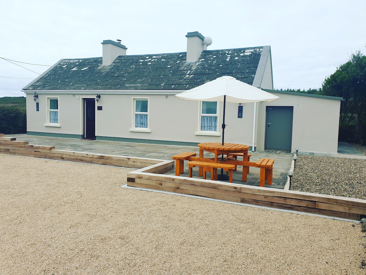 A traditional stone cottage features a light exterior, with a pitched roof and multiple windows. In front, a wooden picnic table is shaded by a large umbrella, surrounded by a gravel area. The landscape beyond highlights the peaceful rural setting.