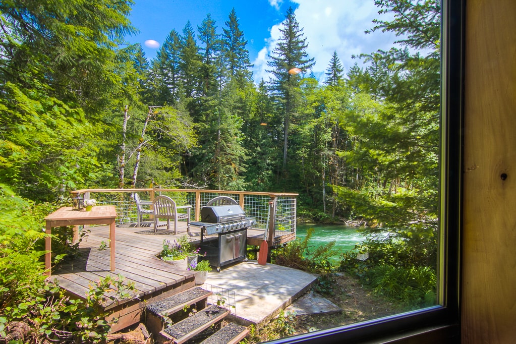 A deck area surrounded by trees is featured, with a grill placed centrally on a wooden surface. Nearby, seating is available, providing a space to relax and enjoy the views of the lush forest and the river flowing beyond.