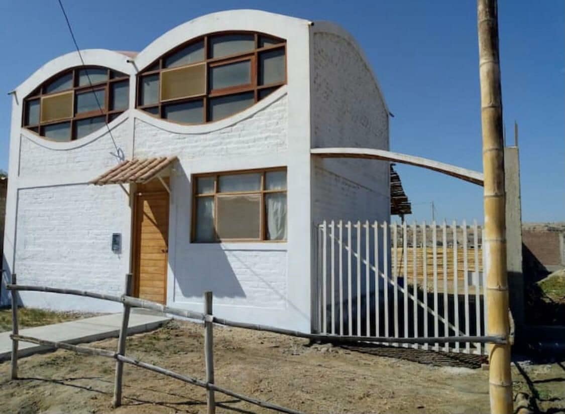 A modern, two-story building is featured with distinct arched window designs. A wooden door welcomes guests, while a metal gate provides secured entry. A gravel pathway leads to the entrance, surrounded by a simple wooden fence.