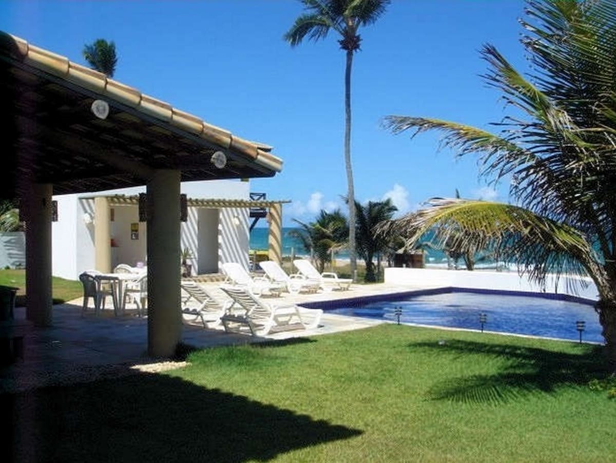 A private pool is surrounded by well-maintained green grass and palm trees. Loungers are arranged near the pool, offering a place to relax. A covered patio area provides shade, with the ocean visible in the distance under a clear blue sky.