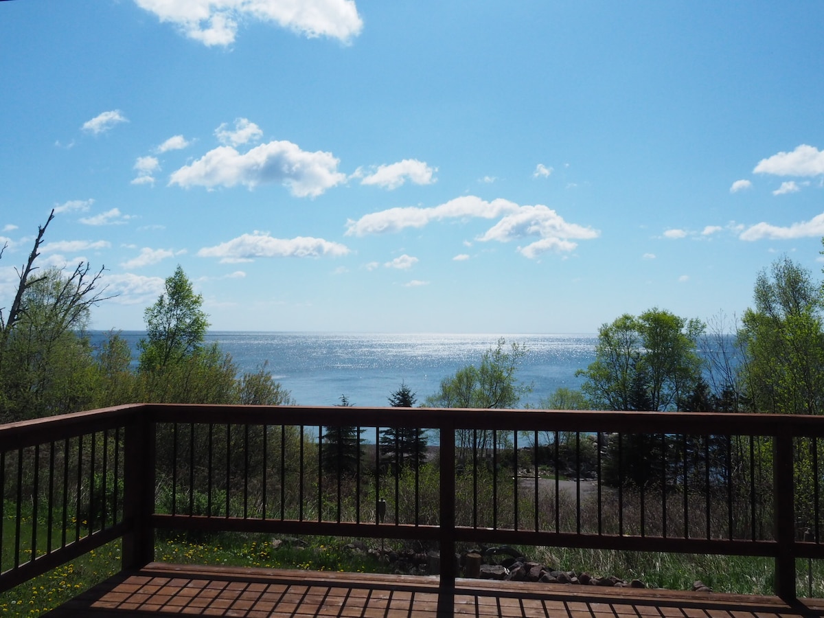 A spacious deck overlooks Lake Superior, where the sun reflects off the water's surface. Lush greenery and trees frame the view, creating a serene backdrop under a bright blue sky with scattered clouds.