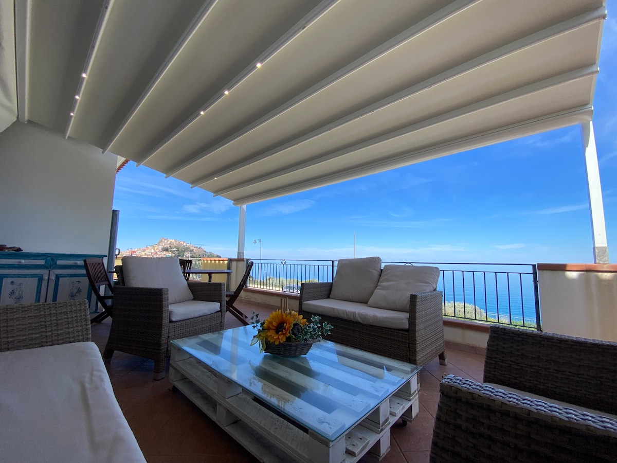 An open veranda features comfortable seating with light-colored cushions, a coffee table with a floral arrangement, and views of the sea and distant landscape. A retractable awning provides shade, while natural light brightens the space, creating an ideal area for relaxation.