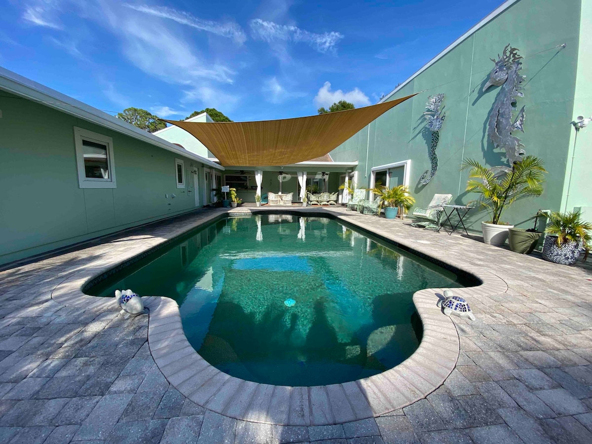 A saltwater pool is set in a spacious outdoor area, surrounded by stone paving. Sunlight reflects off the water, with a shade sail overhead providing relief from direct sunlight. The green walls of the home and decorative seahorse accents enhance the tranquil setting.