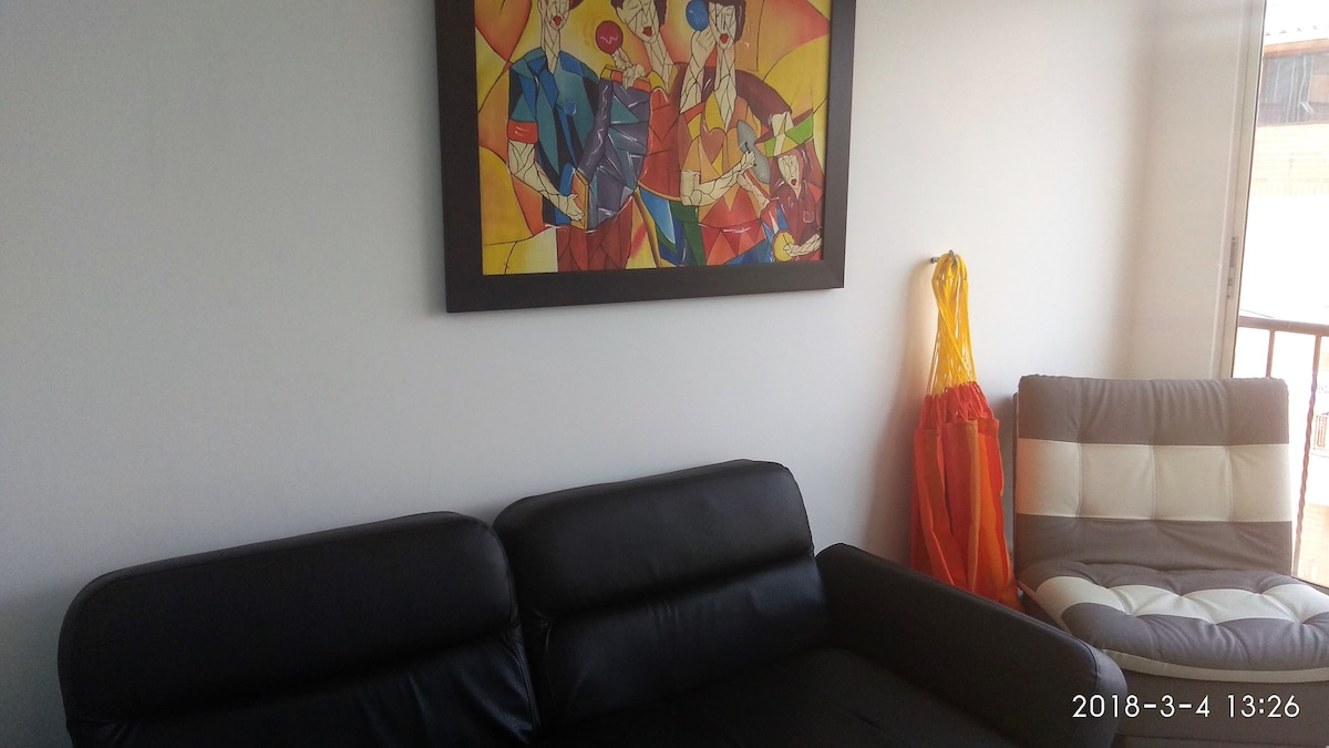 A modern living area features a dark leather sofa paired with a light leather armchair. A colorful mural hangs on the wall, adding a vibrant touch. Natural light flows through the nearby window, enhancing the space's openness.