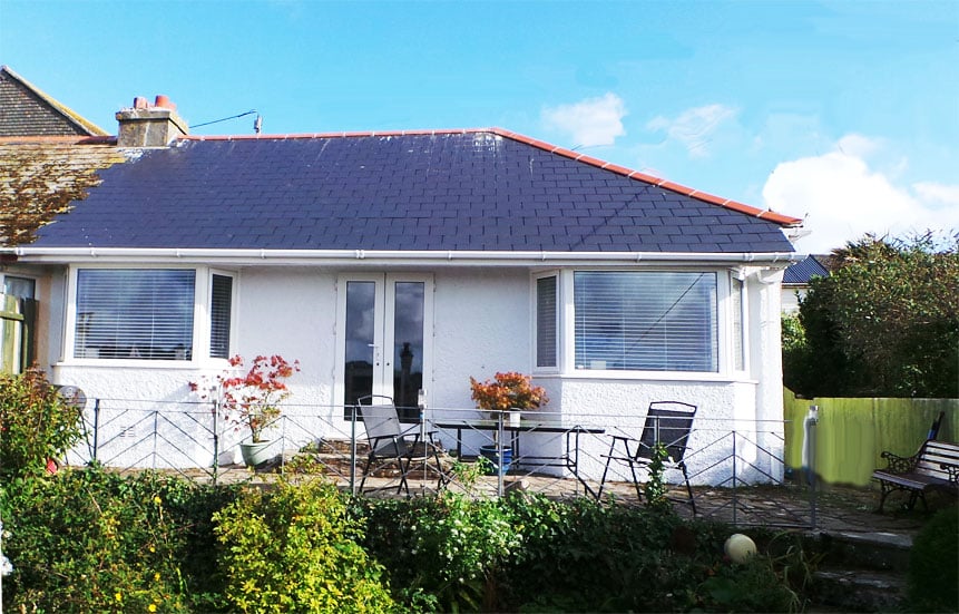 The bungalow's exterior is shown, featuring a sloped, blue roof and large glass doors that open onto a patio. Two chairs and a small table are placed in front of the house, surrounded by a well-maintained garden with colorful foliage.