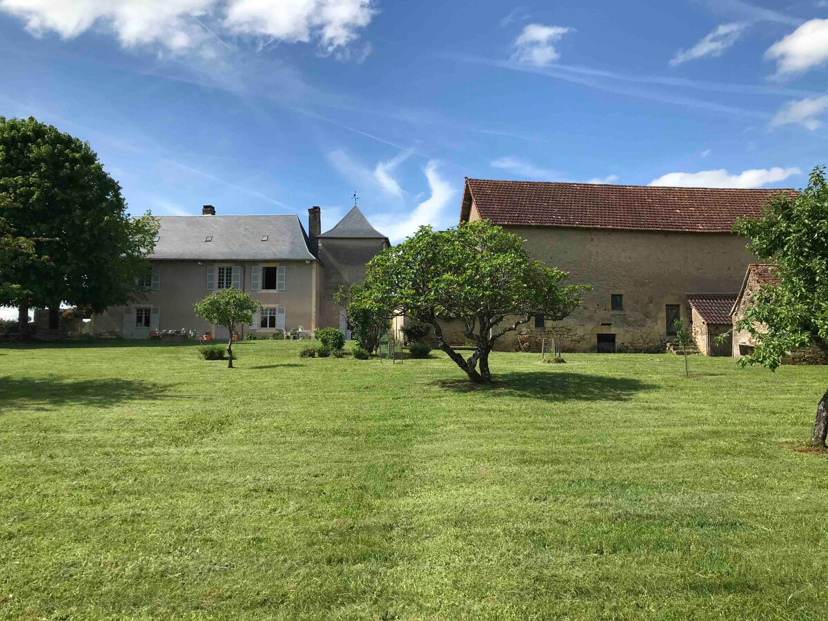 A spacious green lawn is surrounded by two large buildings: a charming house with a slate roof and a rustic stone structure. A mature tree provides shade, and the clear blue sky adds to the serene atmosphere of the property.