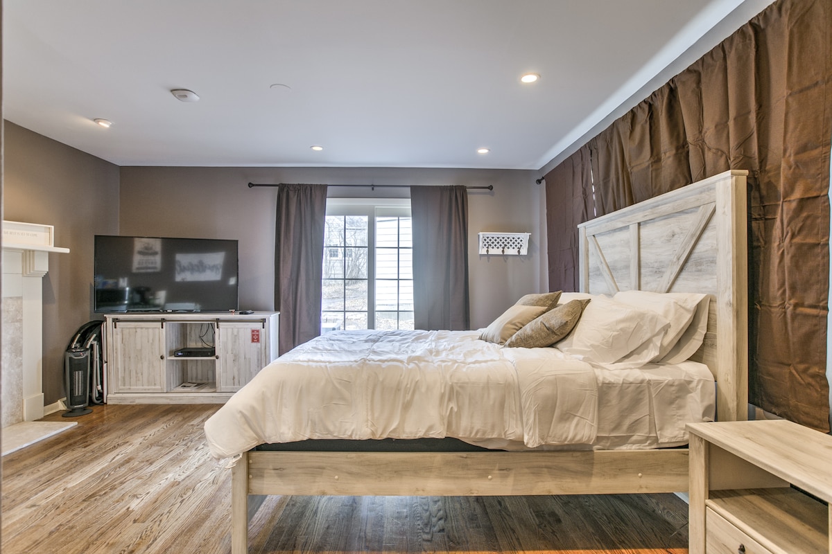 A spacious bedroom features a light-colored wooden bed with white bedding. Nearby, a television is mounted on a white media console. Natural light enters through a window, accented by dark curtains. A decorative fireplace adds warmth to the room's neutral tones.
