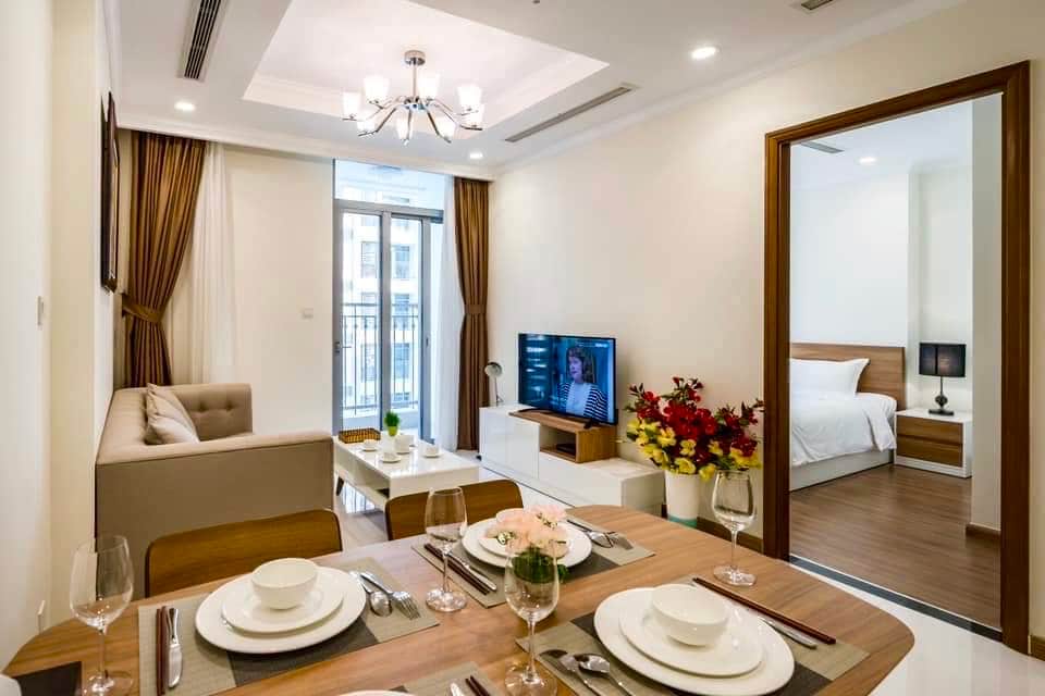 An open concept living area showcases a dining table set for a meal, complemented by a comfortable sofa and a television. Soft drapes frame the window allowing natural light, while a doorway leads to a bedroom featuring a neatly made bed, enhancing the space's overall layout.