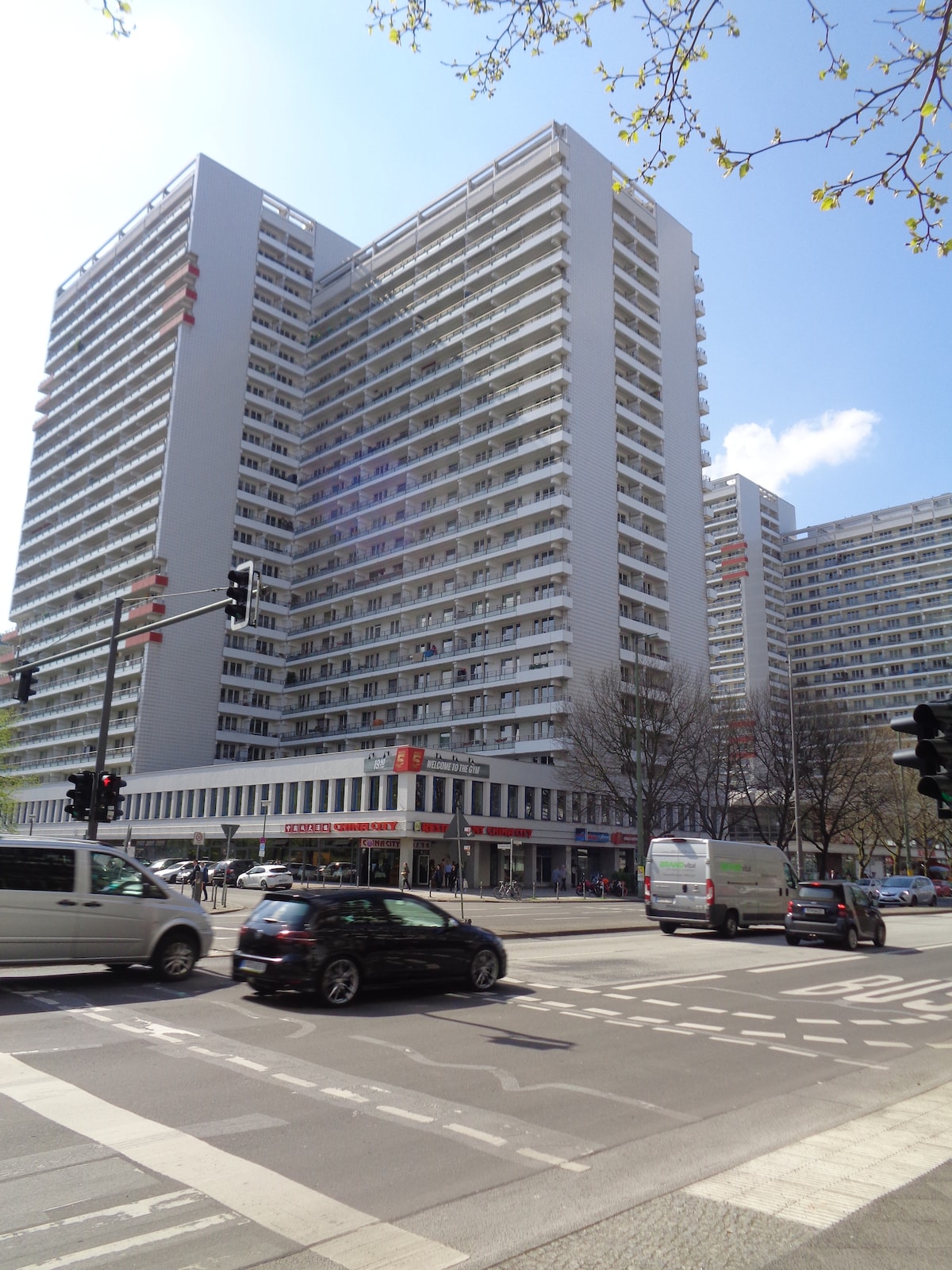 A modern high-rise building is shown, featuring multiple floors with a façade of large windows and balconies. The image includes a street view with passing vehicles and trees lining the road, indicating an urban environment in Berlin.