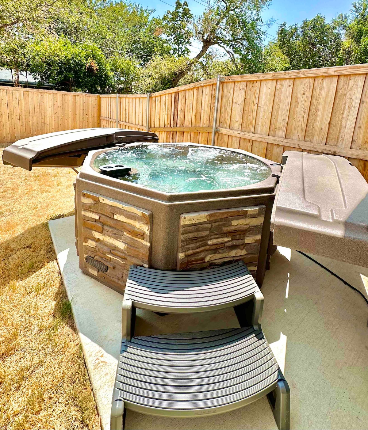 A hot tub is positioned on a concrete patio, surrounded by a wooden fence that enhances privacy. Steps lead up to the hot tub, which features a sturdy lid. Sunlight reflects off the water's surface, creating a serene atmosphere.