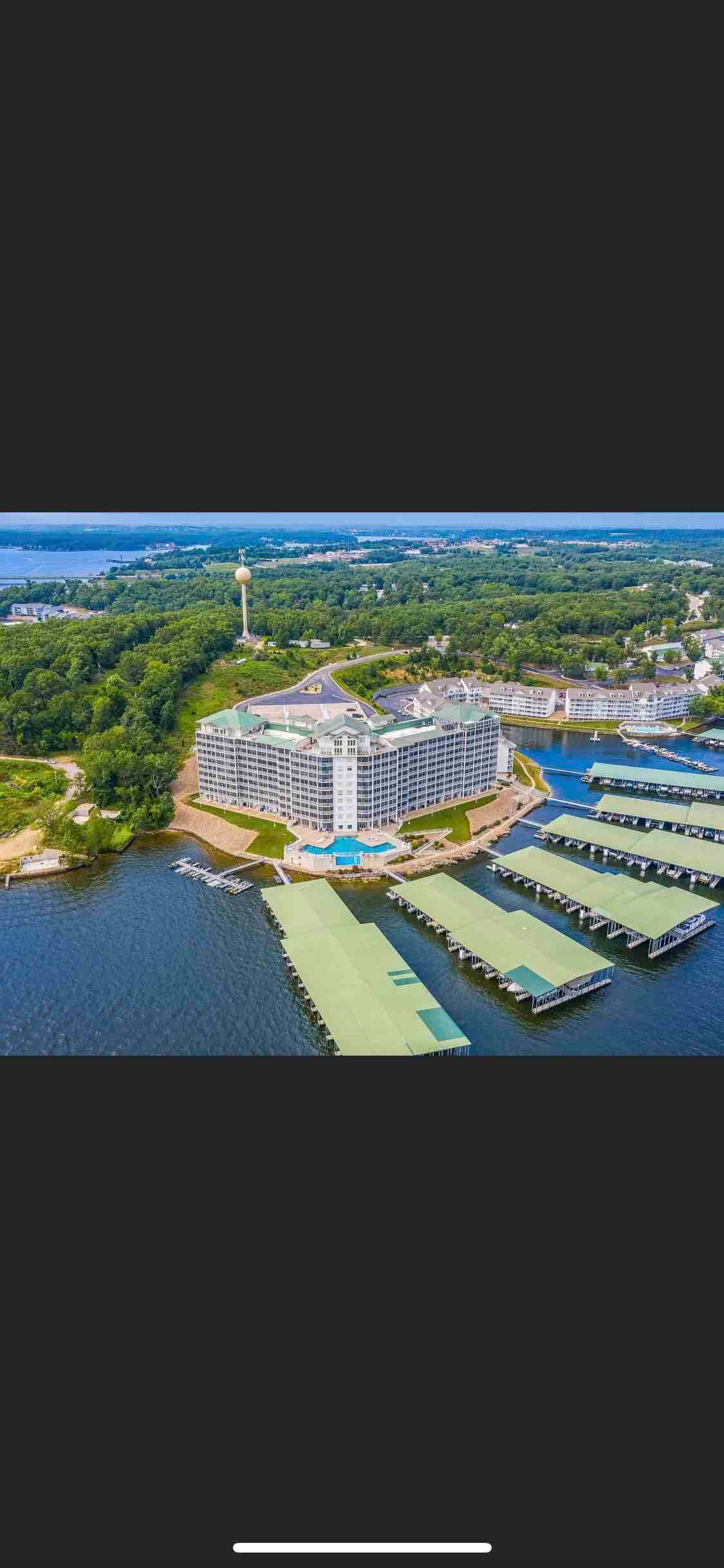 An aerial view showcases the iconic condo building nestled against the lake, framed by lush greenery. Marinas with covered boat docks extend along the waterâs edge, completing the serene lakeside setting.