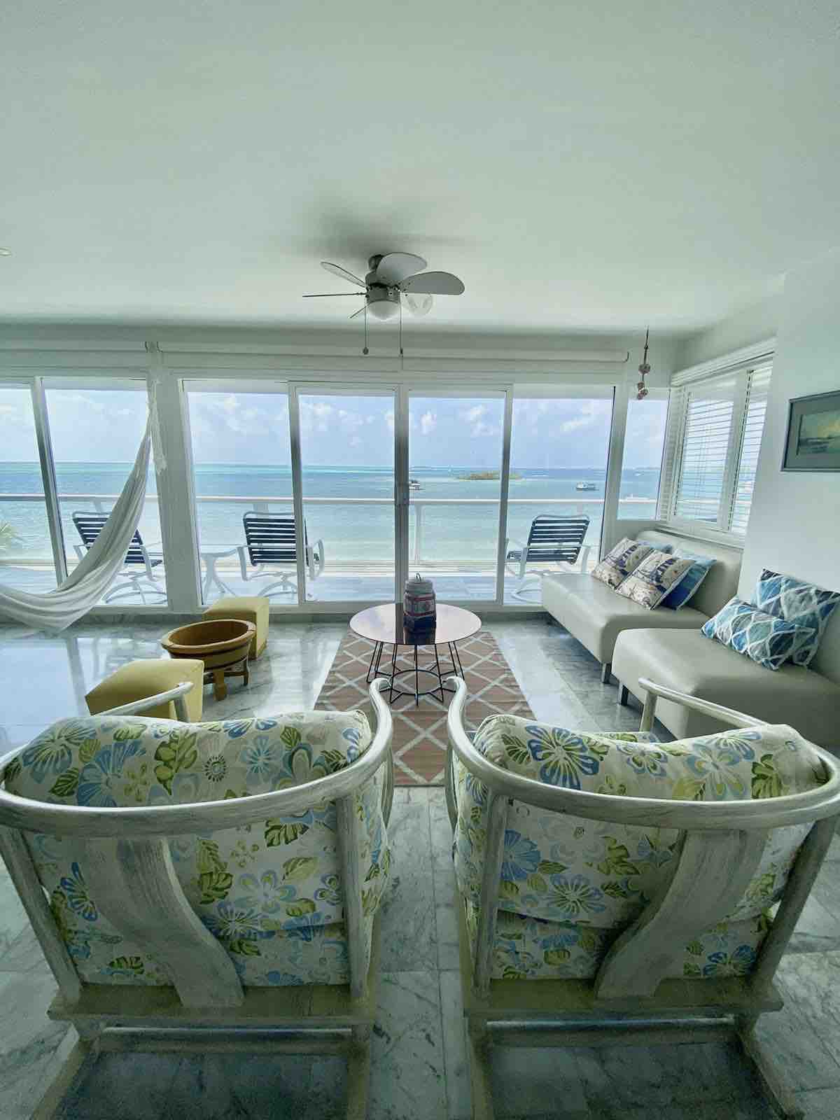 A bright and airy living space features large windows that provide expansive views of the ocean. Two chairs with floral upholstery are positioned around a small table, complemented by a relaxing hammock. A sofa with coastal-themed cushions offers additional seating beside a rug that adds warmth.