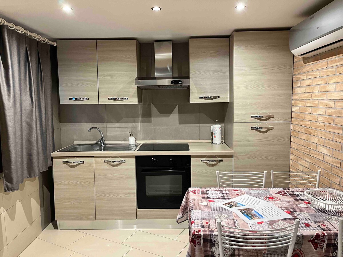 The compact kitchen features sleek cabinetry in a light wood finish, equipped with essential appliances including a stove and oven. A round dining table is set with white chairs, complemented by a checkered tablecloth. Soft lighting creates a warm atmosphere, enhancing the inviting space.