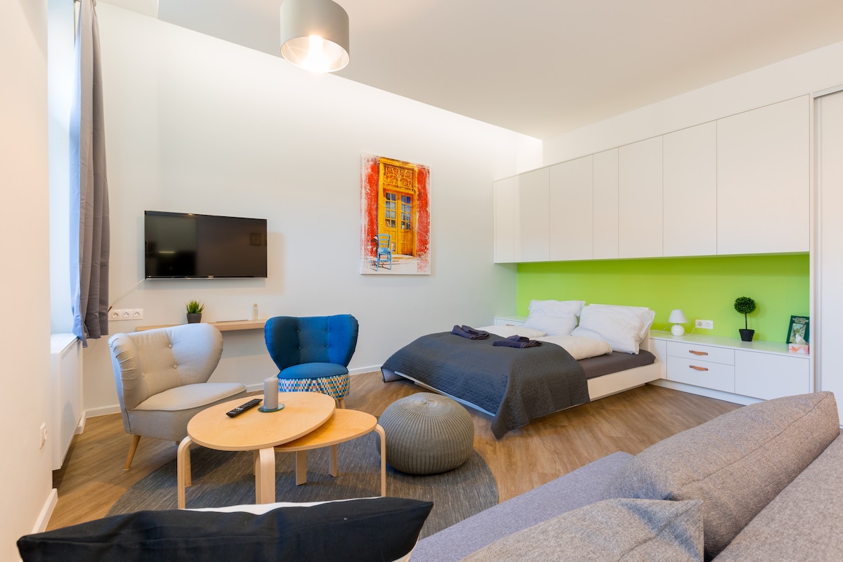 A studio space featuring a large double bed with white bedding and a blue couch. Two light-colored chairs and a round wooden table are arranged in front of the couch. A flat-screen TV is mounted on the wall, and vibrant artwork complements the room's lively design.