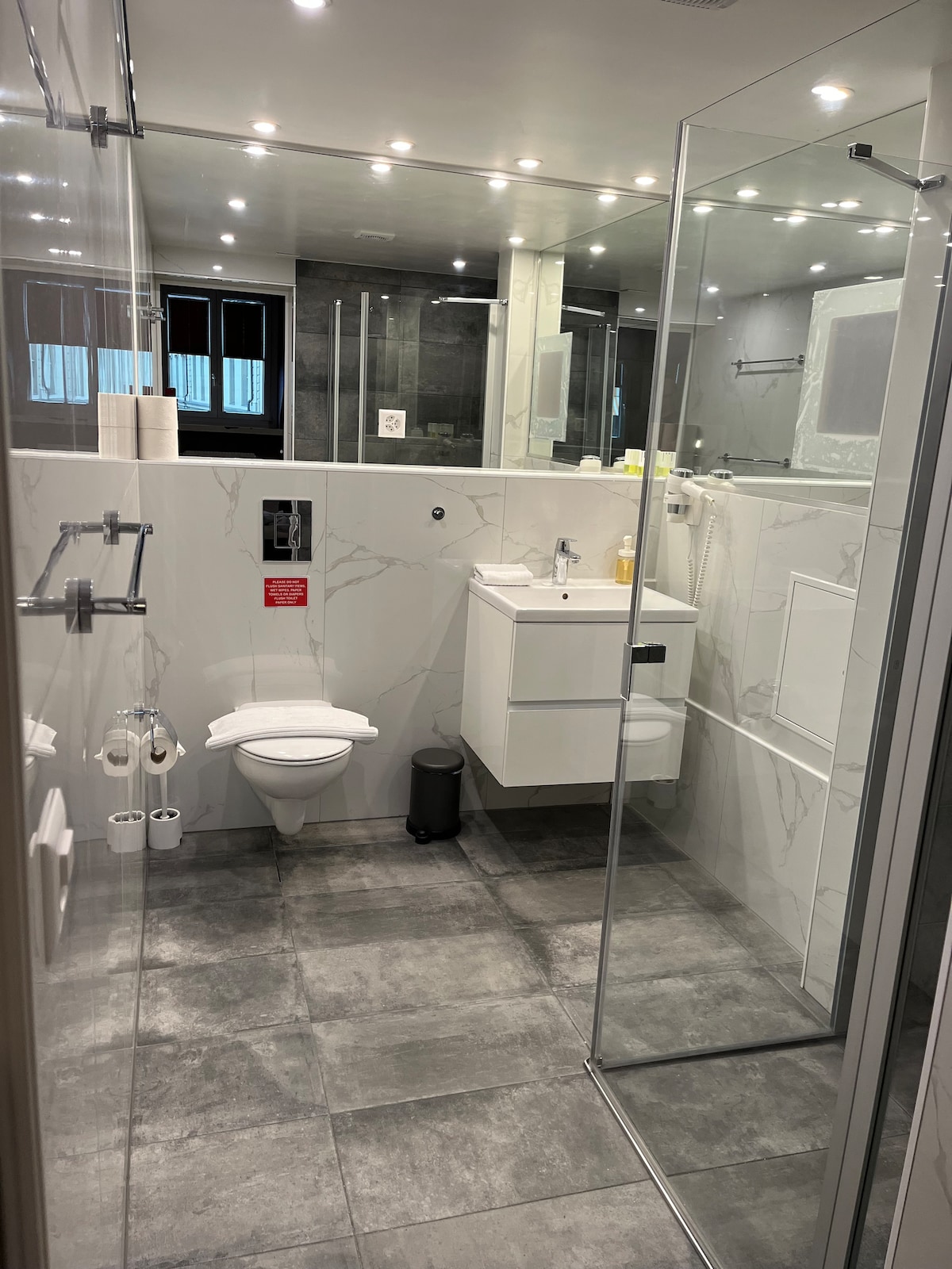 A modern bathroom is presented with tiled flooring in shades of gray. The space features a walk-in shower with glass panels, a wall-mounted sink with storage below, and a toilet. Light fixtures offer illumination, and towels are neatly arranged on a rack.