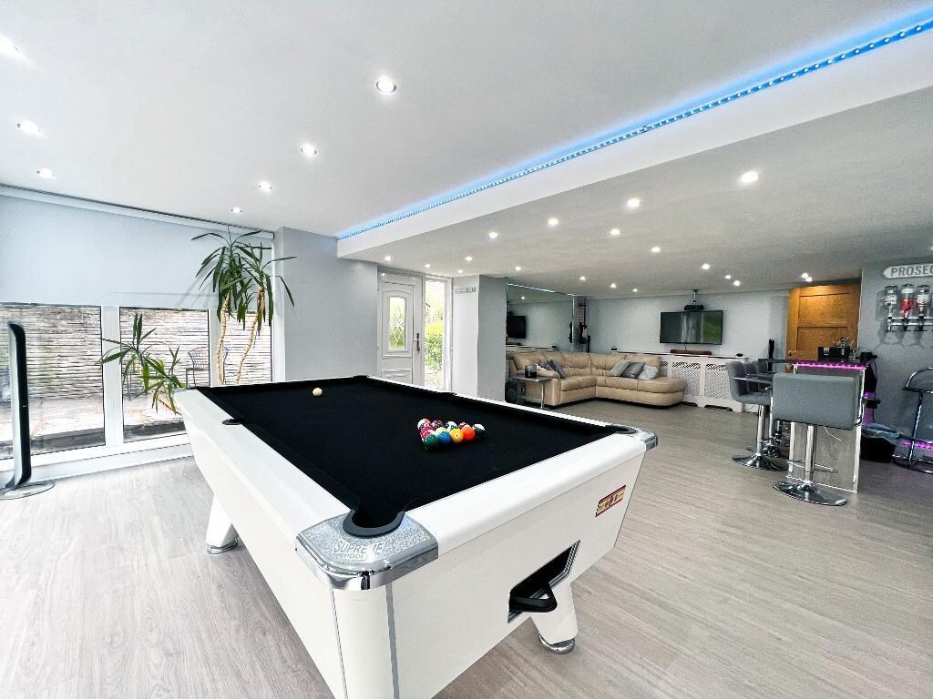 A bright and modern game room features a sleek black pool table at the center, with a set of colorful balls arranged for play. Large windows allow natural light to fill the space, illuminating the contemporary furniture and bar area in the background.