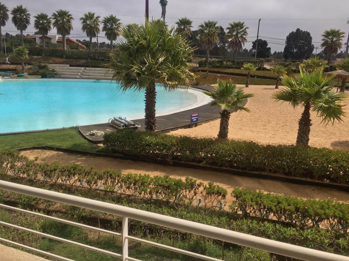 A tranquil scene of a lagoon-style pool surrounded by manicured gardens and palm trees. A sandy area is visible nearby, providing a beach-like atmosphere. Pathways line the space, allowing easy access to the water and sandy zones.