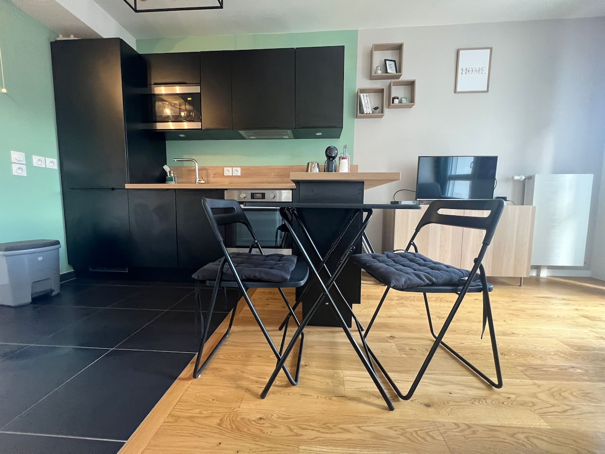A modern kitchen area features black cabinets, a wood-accented countertop, and stainless steel appliances. A small dining table with two folding chairs is positioned in front of the kitchen. A television and additional furnishings complement the open space, illuminated by natural light.