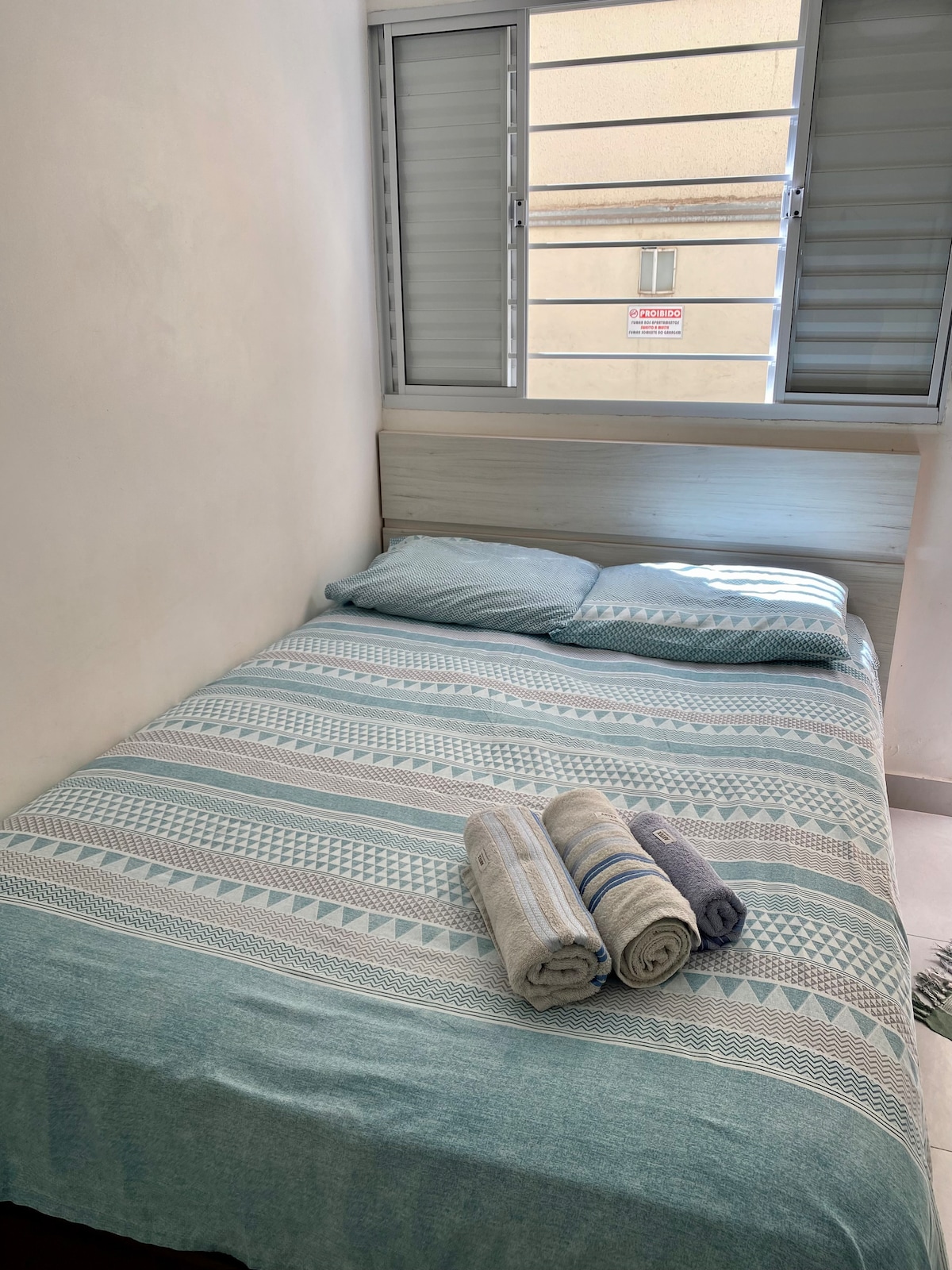 A comfortable sleeping area is presented with a spacious bed topped with a light blue patterned blanket. Fresh towels are neatly arranged at the foot of the bed. Natural light filters through a set of partially open windows with simple horizontal blinds.