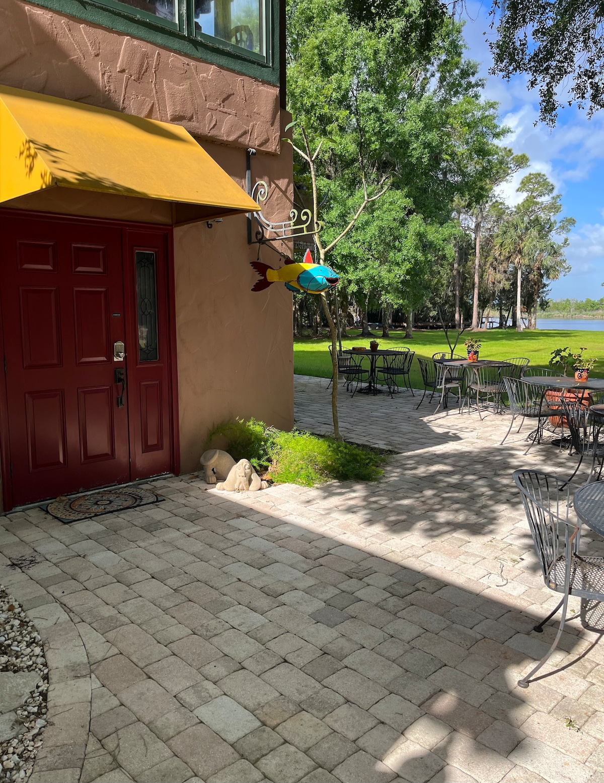 A welcoming entrance features a bright red door beneath a yellow awning. A pathway made of pavers leads to several outdoor seating arrangements, with black metal tables and chairs. Lush green grass and trees are visible in the background, offering a serene atmosphere.