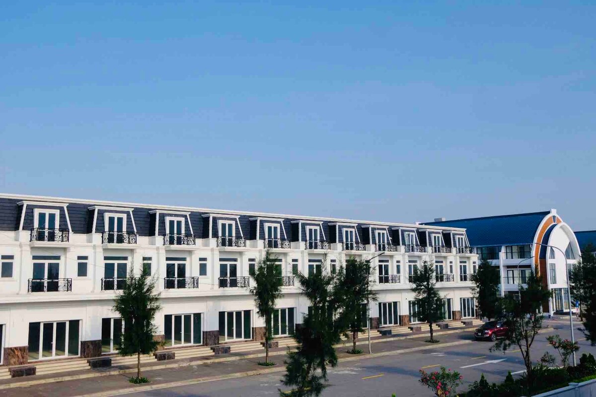 A modern building features a series of windows and balconies along its façade, surrounded by landscaped greenery. A separate structure with a curved roof is visible in the background, contributing to the inviting layout that accommodates various guests.
