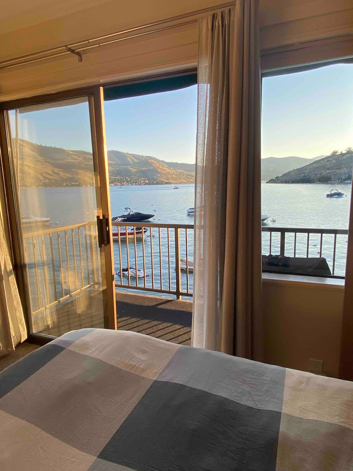 A sliding glass door opens to stunning lake views, with boats gently floating on the water. Natural light spills into the space, highlighting the soft colors of the bedding. The surrounding landscape features hills and greenery, creating a serene backdrop.
