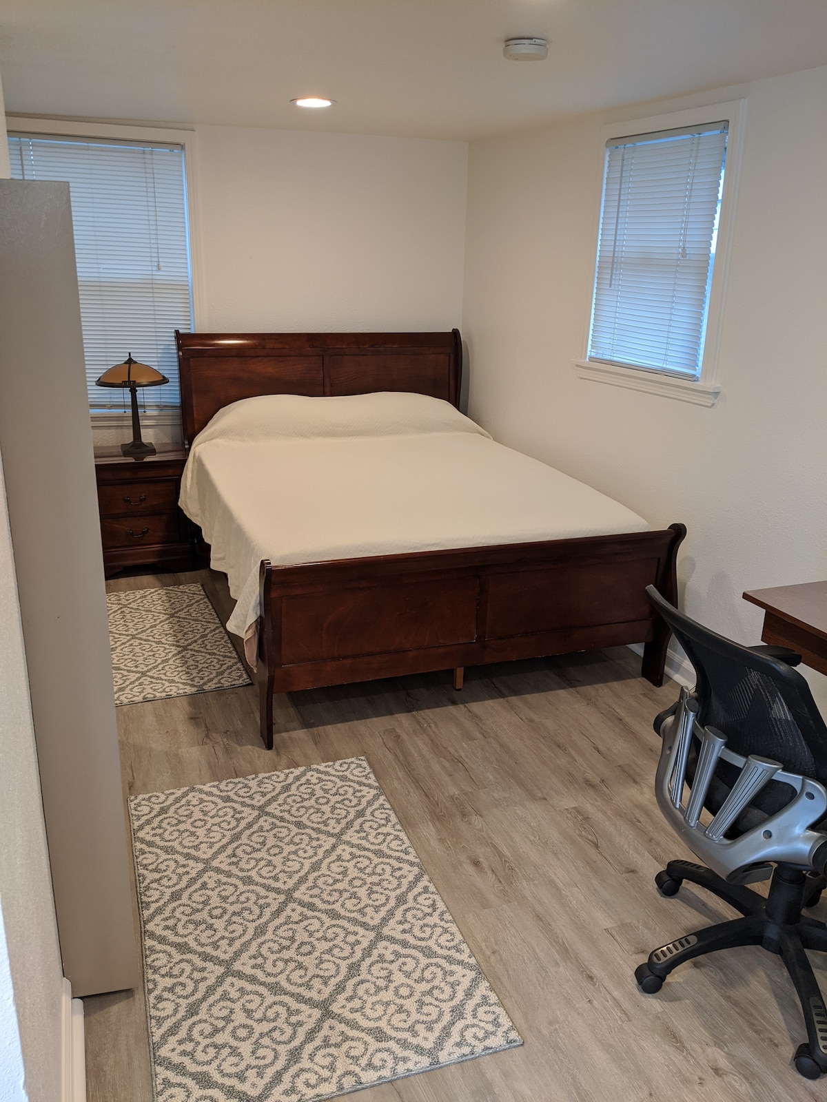 A cozy bedroom features a queen bed with a simple white coverlet and a wooden headboard. A bedside lamp and a small nightstand are present alongside the bed. A chair and desk are positioned nearby, while patterned rugs enhance the floor space.