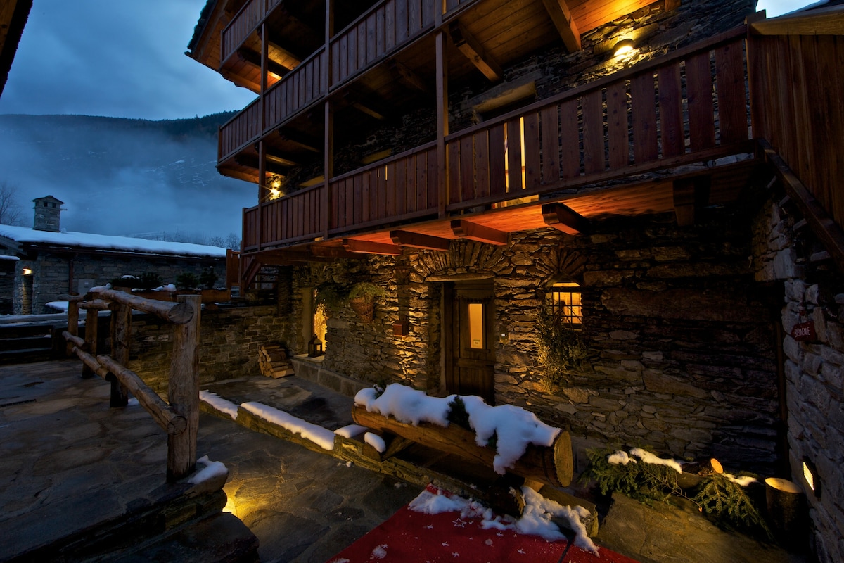 The exterior of a charming mountain residence features stone walls and wooden balconies, lit warmly in the twilight. Snow blankets the ground, and soft lighting enhances the inviting entrance, which is complemented by rustic wooden accents and a welcoming red carpet.