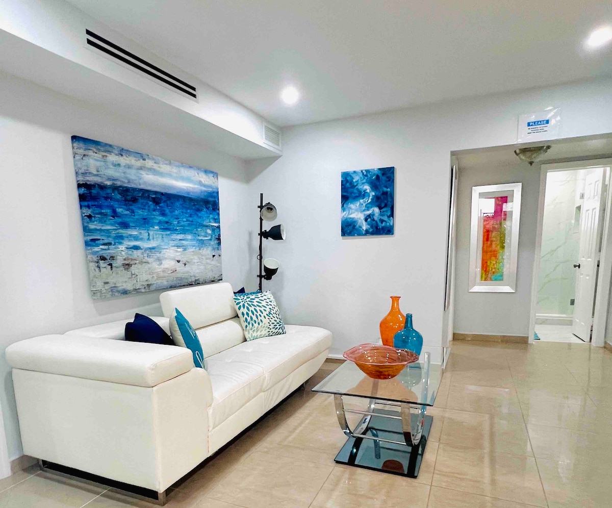 A bright living area features a modern white sectional sofa with colorful throw pillows, positioned near a glass coffee table. Abstract artwork in shades of blue adorns the walls, complemented by decorative glass vases in vibrant colors. An inviting atmosphere is created by ample lighting.