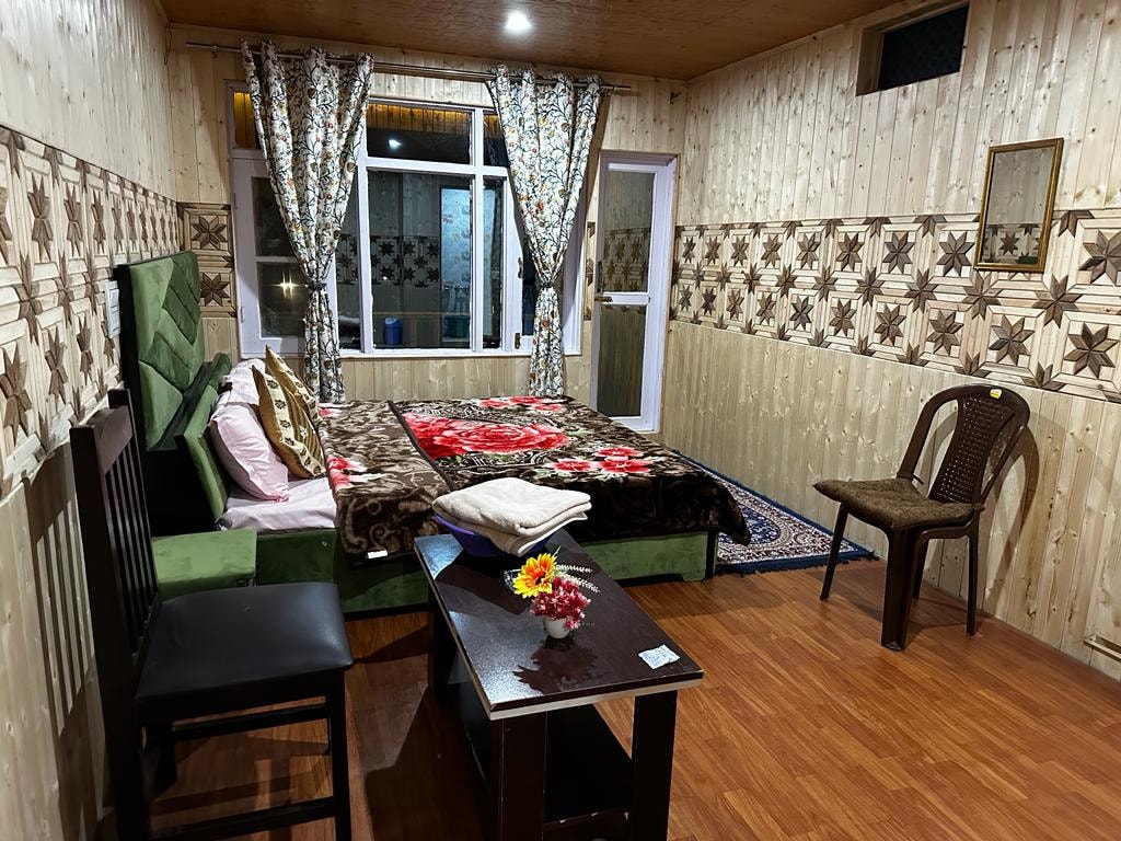 A cozy bedroom is showcased with wooden paneling and decorative wall patterns. A spacious bed, adorned with a patterned blanket, is complemented by light curtains draping from two windows. A simple table with a small flower vase and a wooden chair complete the inviting space.