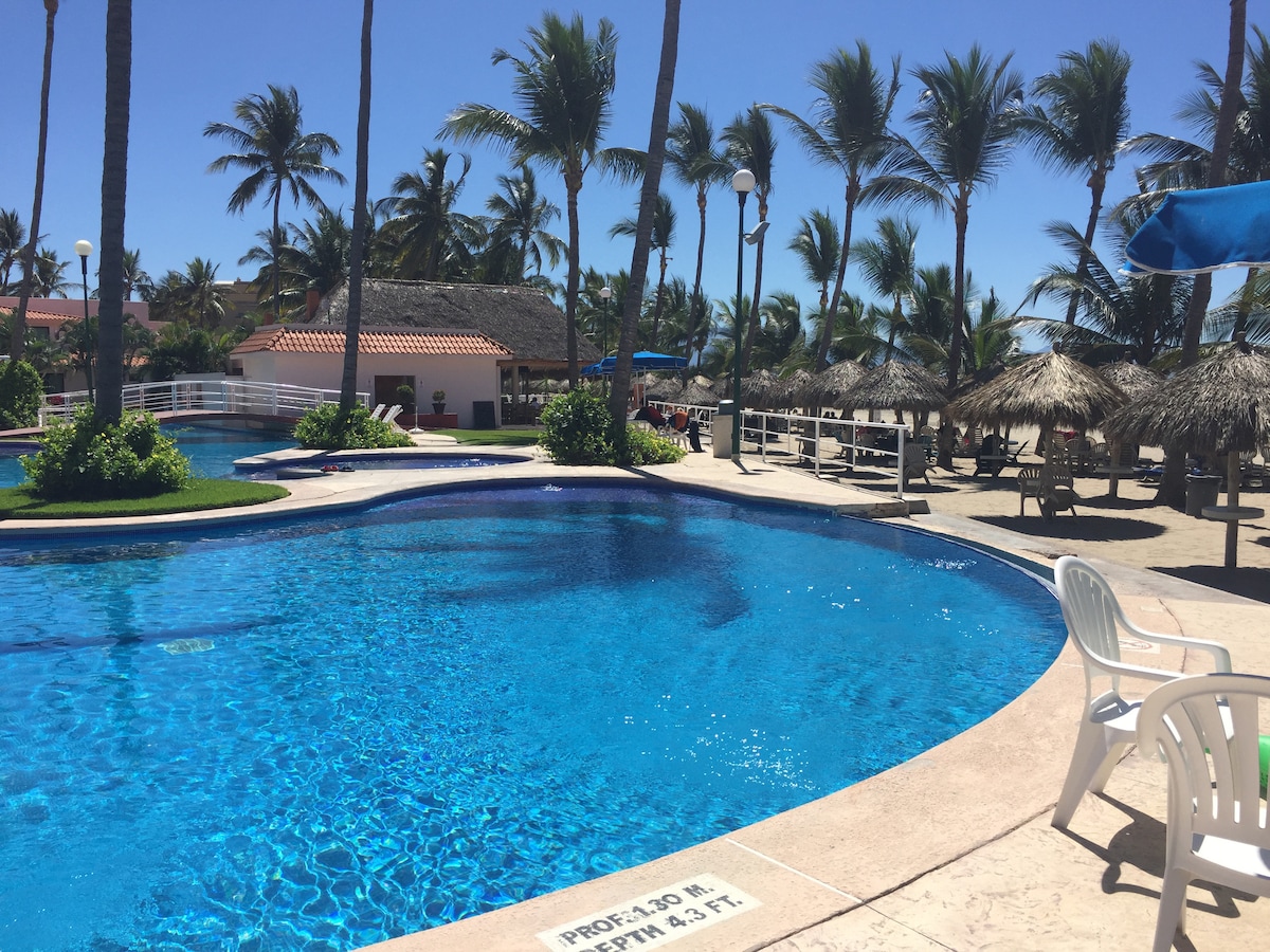 A sparkling blue swimming pool is surrounded by palm trees, providing a tropical ambiance. Lounge chairs are positioned under shaded umbrellas, and thatched-roof structures can be seen in the background, offering seating areas for relaxation.