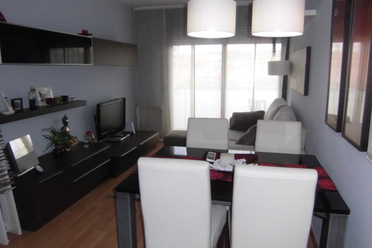 A modern living area is presented with a comfortable gray sofa and a sleek black TV stand. A glass dining table is surrounded by four white chairs, while natural light filters through the large windows, enhancing the light blue wall and contemporary decor.