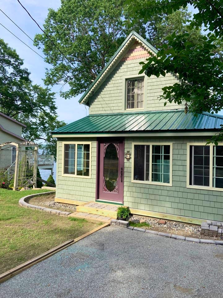 A charming lakefront cottage is displayed with a green exterior and a steeply pitched roof. The entrance features a deep purple door, surrounded by large windows. A well-maintained lawn leads up to the cottage, with visible glimpses of the lake in the background.