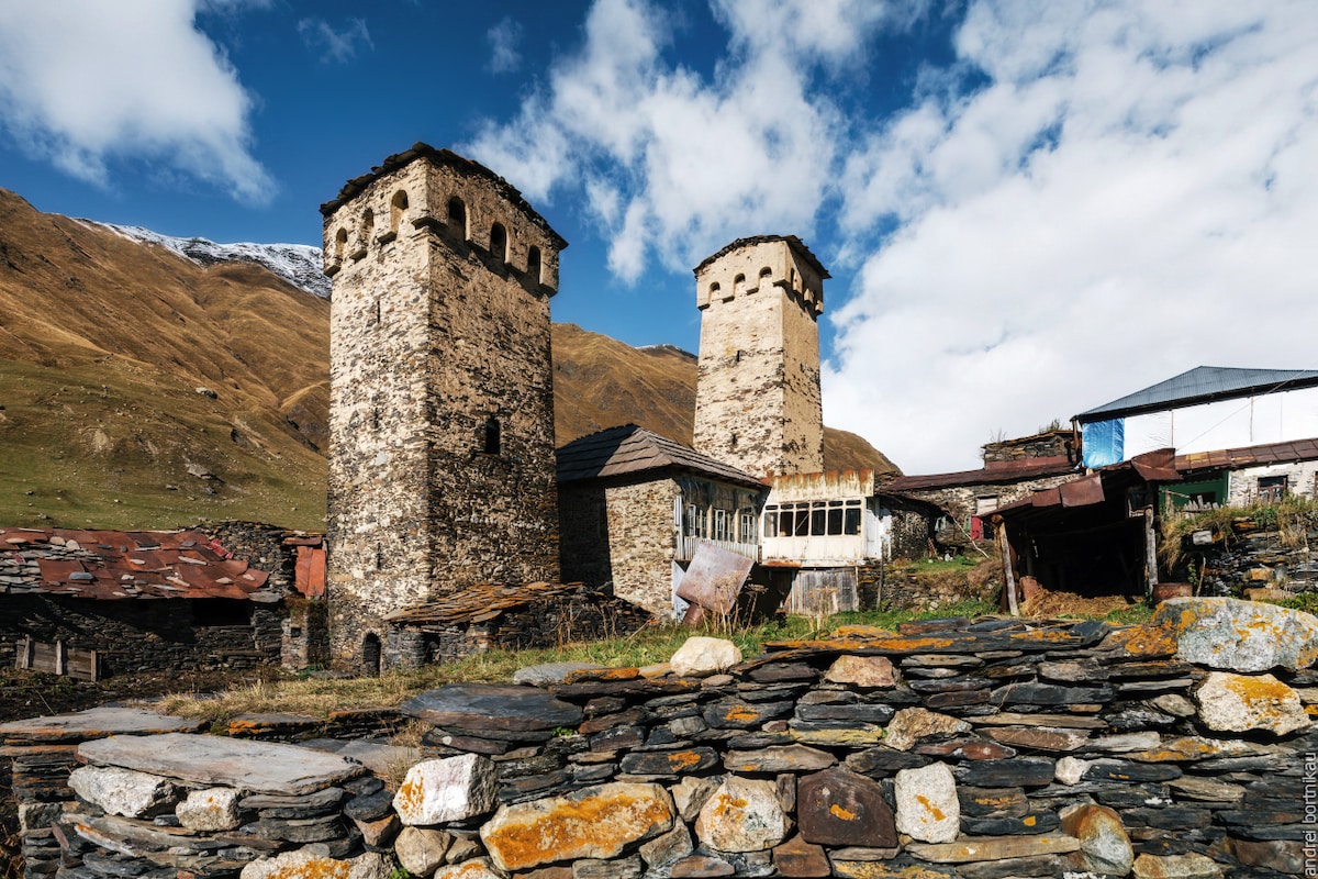 Two stone towers, characterized by their rounded tops, rise amidst a mountainous landscape. A mix of traditional and modern buildings can be seen, with a backdrop of softly sloping hills and a partly cloudy sky.