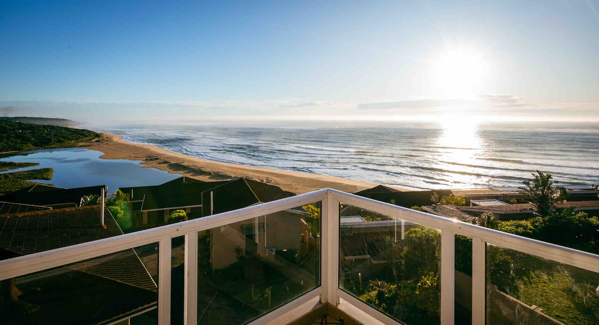A panoramic view from a balcony captures the expansive Indian Ocean and sandy beach under a golden sunrise. The tranquil waves gently roll onto the shore, while lush greenery and rooftops are visible in the foreground, enhancing the serene coastal landscape.
