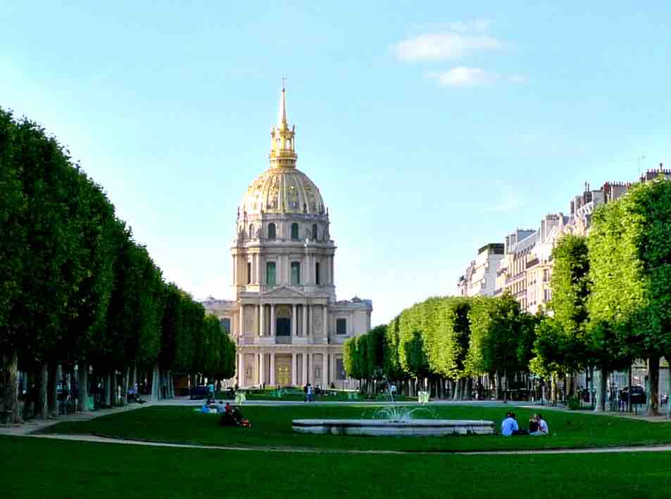 A grand building with a golden dome is surrounded by neatly trimmed trees and manicured green lawns. The scene captures people relaxing on the grass, with a fountain visible in the foreground under a clear blue sky.