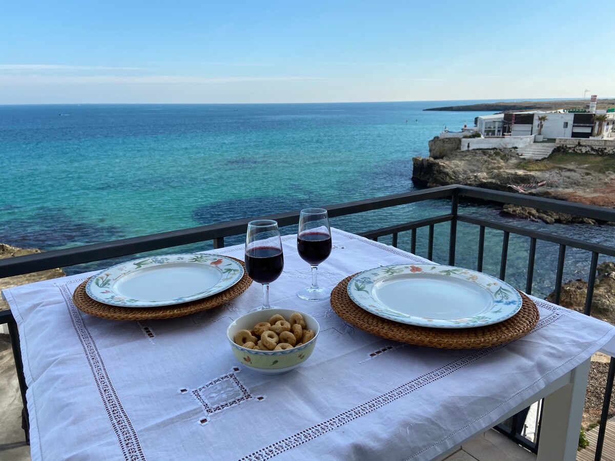 A dining table is set on a balcony overlooking the sea. Two plates are placed beside wine glasses, and a bowl of snacks is centered on the table. The blue water and distant coastline are visible in the background, creating a serene setting.