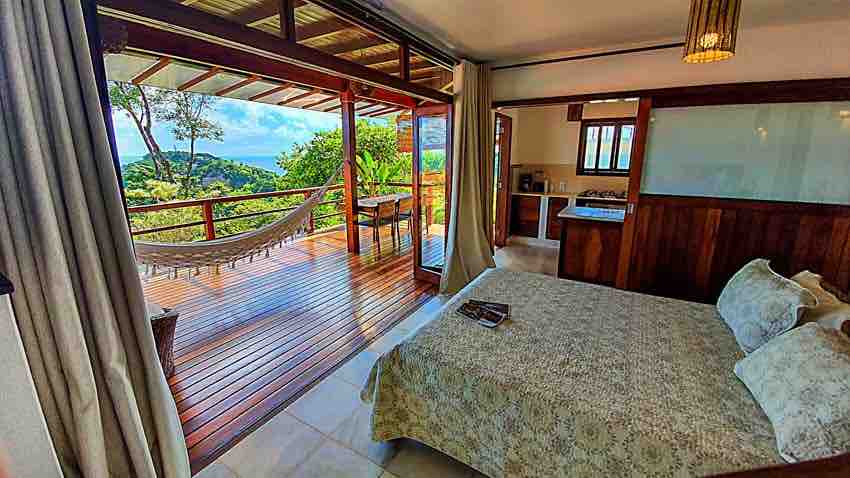 A spacious sleeping area features a light-colored quilt on a large bed, with natural light flowing in from wide sliding doors. A private balcony with a hammock offers scenic views of the surrounding landscape. The kitchenette is visible in the background.