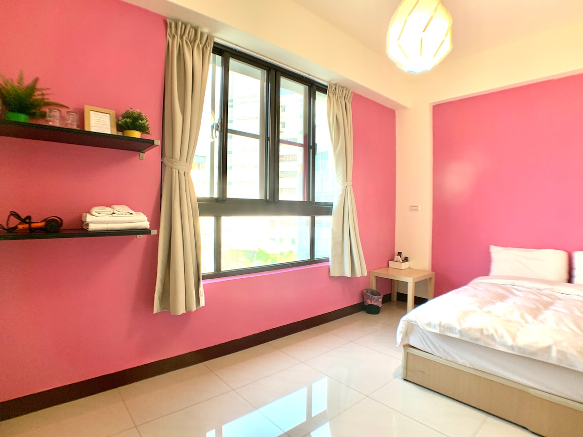 A brightly colored room features a pink accent wall and a spacious bed with white linens. Natural light enters through a large window, illuminating the space. A shelf displays neatly arranged towels and plants, adding a touch of greenery to the vibrant decor.