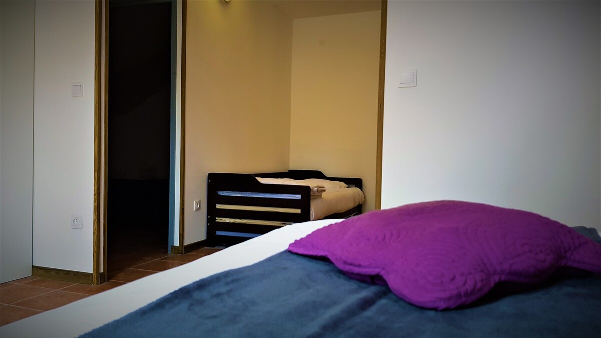 A spacious room is visible, featuring a large double bed in the foreground with a textured purple pillow. A doorway leads to a separate sleeping area containing two single beds, providing additional sleeping accommodations.