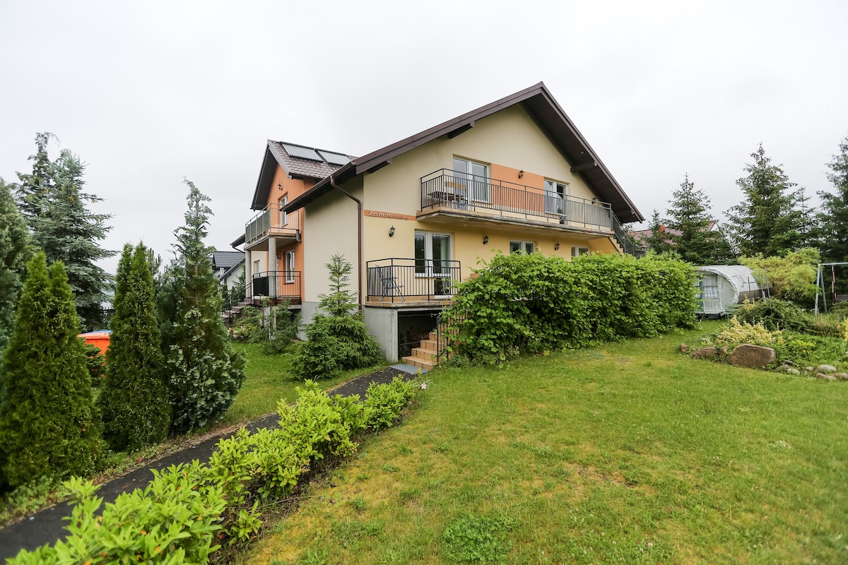 A two-story house is surrounded by lush greenery, featuring multiple balconies with railings and solar panels on the roof. The well-maintained garden includes a pathway leading to the entrance, enhancing the home's inviting exterior.