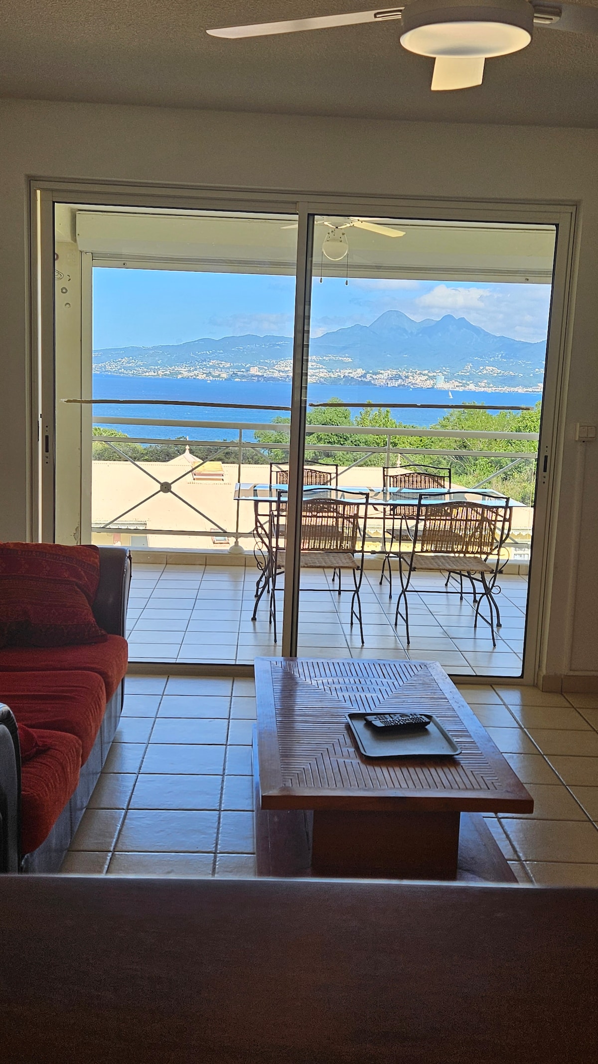A spacious living area is shown, featuring a red sofa facing a large sliding glass door. The door leads to a balcony with outdoor seating and a table, offering a panoramic view of mountains and the bay beyond.