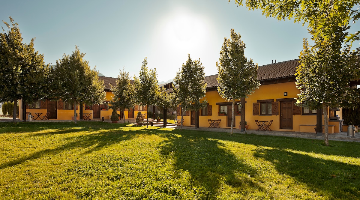A vibrant garden area features neatly trimmed grass and several trees. Two buildings, painted in warm yellow, are arranged with outdoor seating areas equipped with small tables and chairs. Soft sunlight bathes the setting, creating a serene environment.