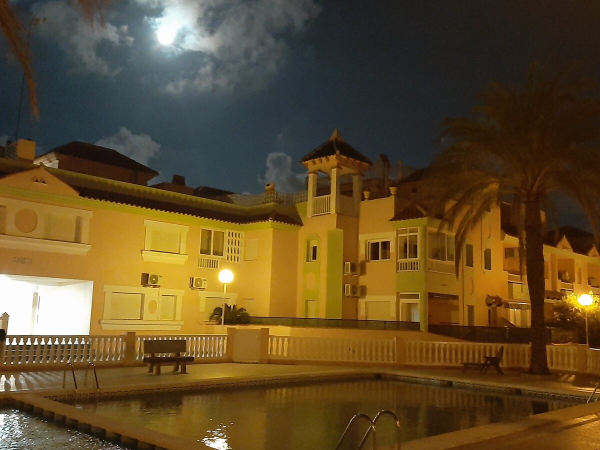 The image captures a tranquil evening scene featuring a well-lit community pool surrounded by palm trees. The illuminated buildings, with light reflecting off their pastel facades, suggest a peaceful atmosphere. The moon is visible in the sky, enhancing the serene setting.